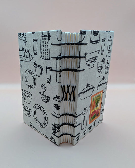 Handmade mini sketchbook with print of utensils, paper is from Nepal. Featuring a beautiful image of veggies on the front. Cold press paper, acid free. Shown standing up.