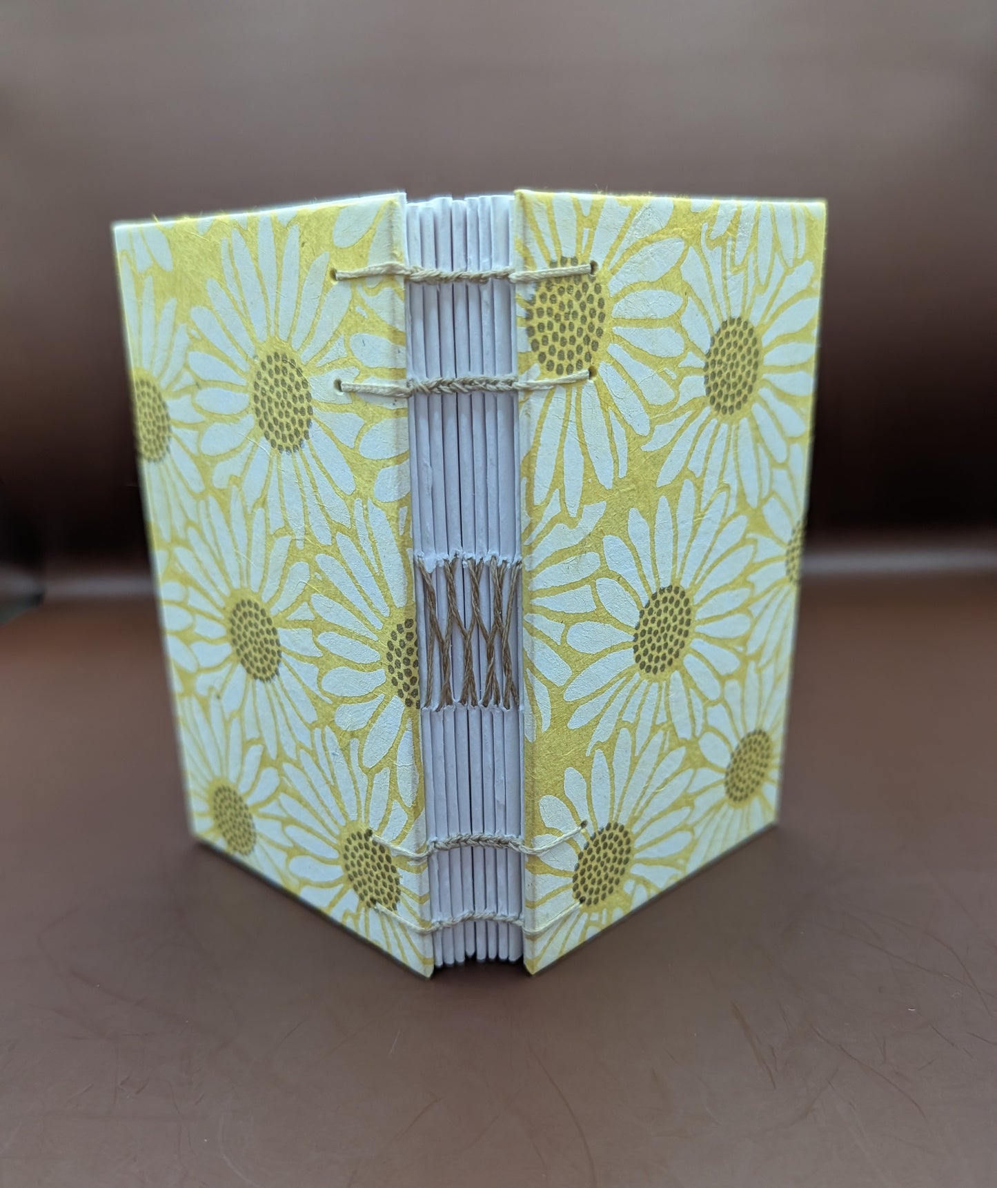 Pocket-sized 3x5" handmade daisies on yellow background pattern. Features 96 pages of cold press paper gorgeous gold French stitch binding, and Nepal paper covers. Shown standing up.