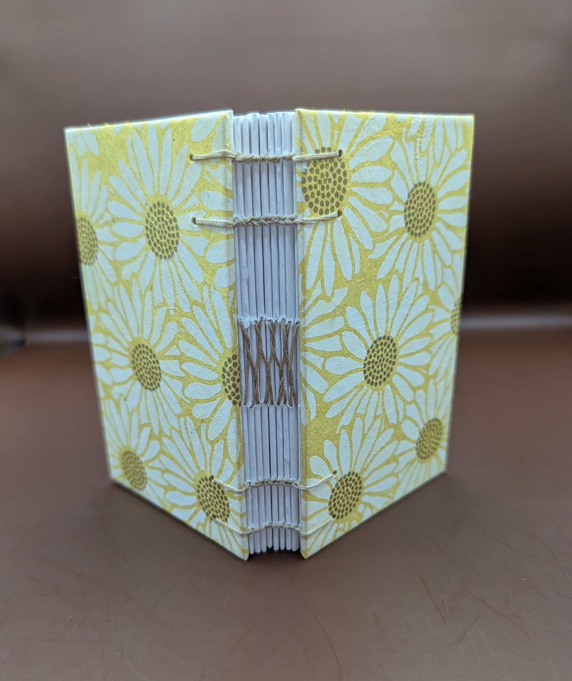 Pocket-sized 3x5" handmade daisies on yellow background pattern. Features 96 pages of cold press paper gorgeous gold French stitch binding, and Nepal paper covers. Shown standing up.