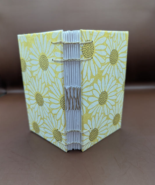 Pocket-sized 3x5" handmade daisies on yellow background pattern. Features 96 pages of cold press paper gorgeous gold French stitch binding, and Nepal paper covers. Shown standing up.