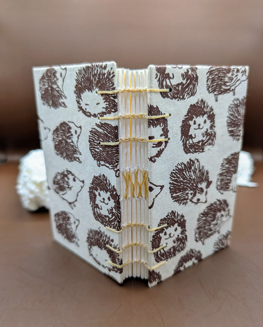 Handmade mini sketchbook featuring a brown hedgehog patter cover and a decorative gold exposed French stitch binding, shown standing open.