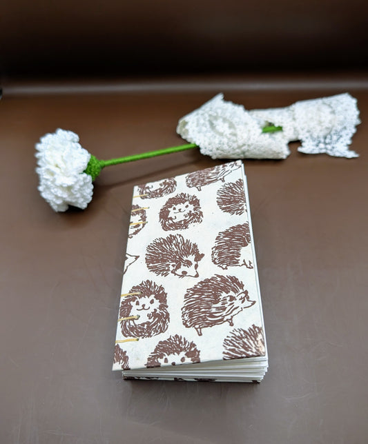 Handmade mini sketchbook featuring a brown hedgehog patter cover and a decorative gold exposed French stitch binding, shown on brown background with a white rose in crochet.