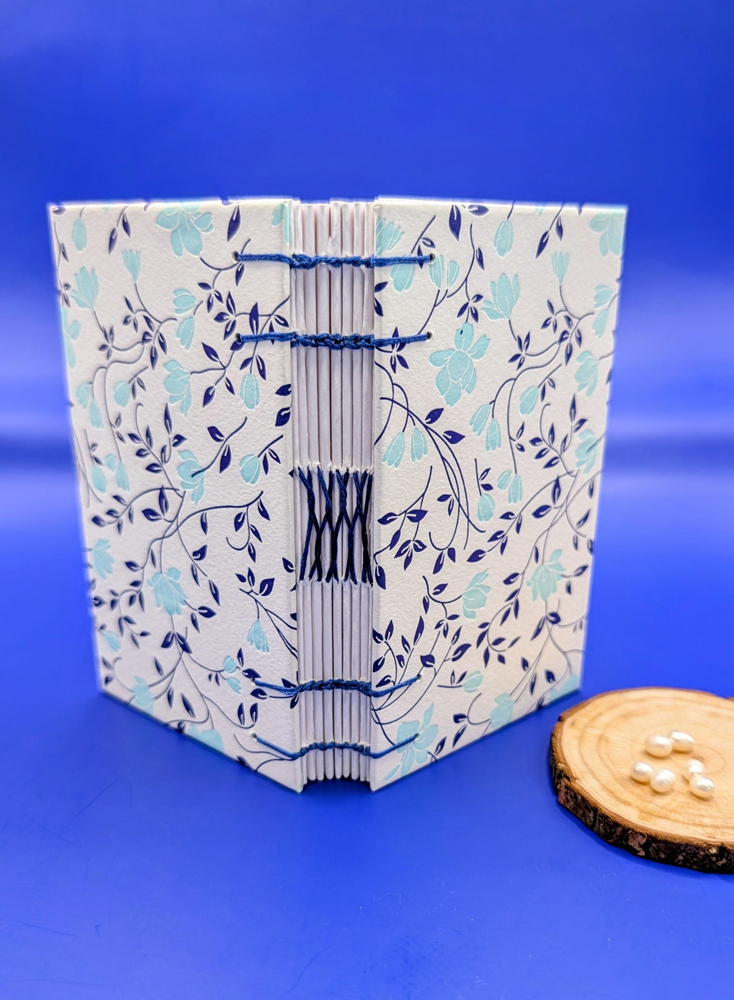 Handmade mini sketchbook featuring turquoise flowers and blue leaves decorative paper from Italy with blue wax thread on an exposed French stitch binding, shown standing open.