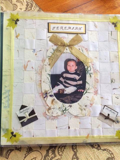 Handcrafted 12x12 baby scrapbook page featuring a central oval photo frame of a baby, woven white paper background, ribbon bow, and small date banners labeled Jeremiah